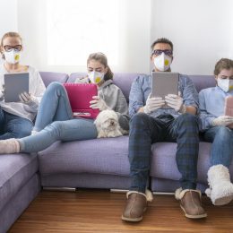 A mother, father, and their two daughters sit on a couch using tablets and devices while wearing face masks.