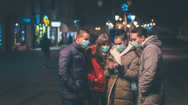 A group of young friends wearing face masks look at a smartphone in one of their hands.