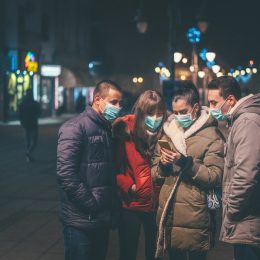 A group of young friends wearing face masks look at a smartphone in one of their hands.