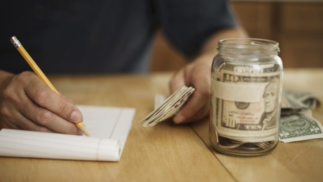 A male writes notes concerning money at the kitchen table. On the table is a pencil, pad of paper, paper currency and a jar fill with cash and coins.