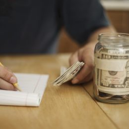 A male writes notes concerning money at the kitchen table. On the table is a pencil, pad of paper, paper currency and a jar fill with cash and coins.
