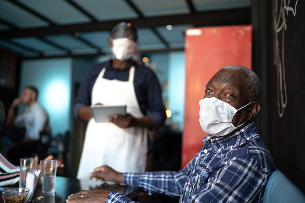 senior black customer while waiter taking order at restaurant amid covid, both are wearing masks