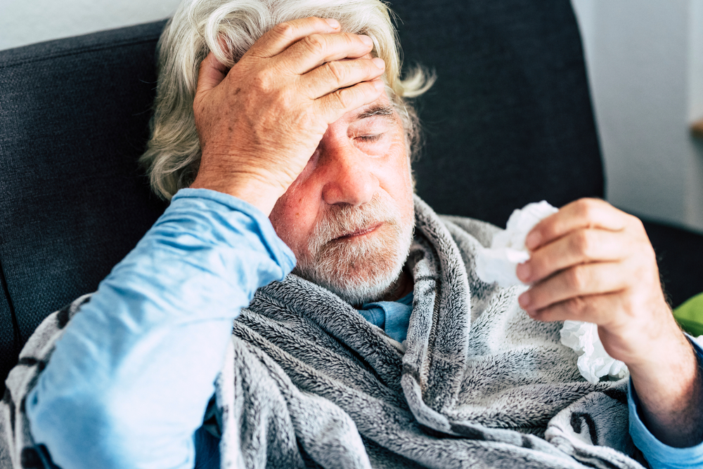A senior man sits on a couch wrapped in a blanket while touching his forehead and feeling for a fever, suffering from COVID symptoms