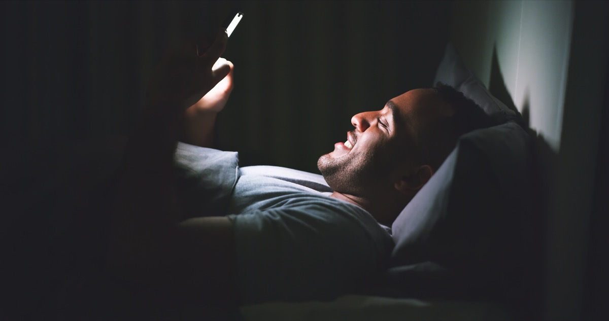 Shot of a cheerful young man using his cellphone while lying in bed late at night