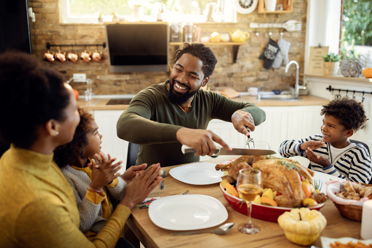Smiling black man having Thanksgiving lunch with his family and carving stuffed turkey at dining table, surrounded by wife and children