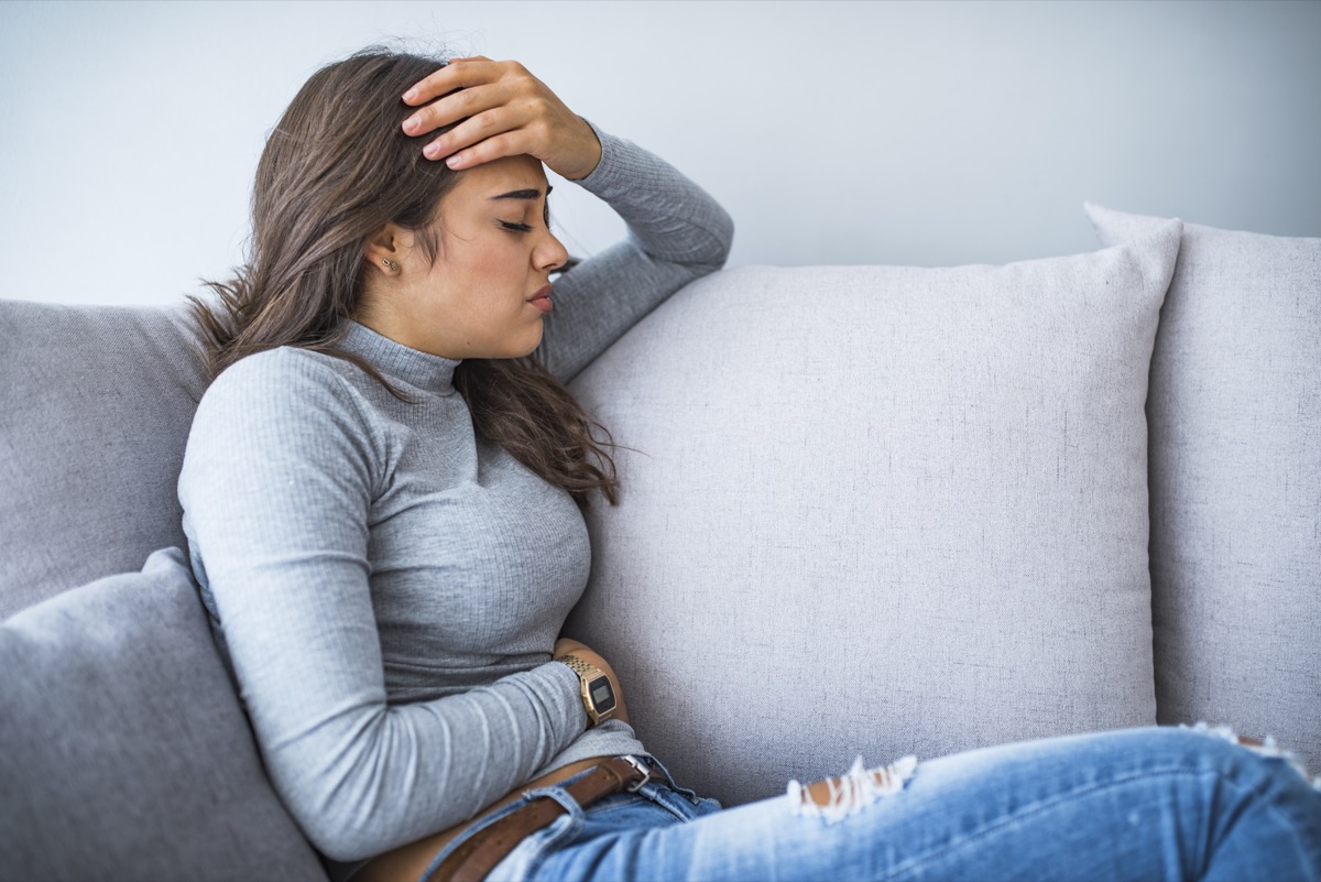 View of young woman suffering from stomachache on sofa at home. Woman sitting on bed and having stomach ache. Young woman suffering from abdominal pain while sitting on sofa at home