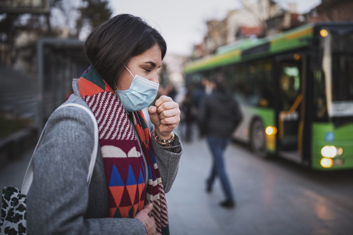Side view of woman wearing a face mask and coughing while standing at bus stop