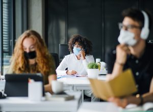 A young woman wearing a face mask sits in an office with coworkers also wearing face masks.