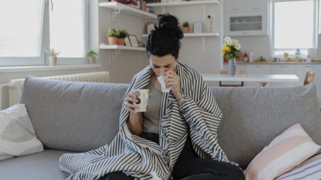 A young woman sits on a couch wrapped in a blanket with COVID symptoms