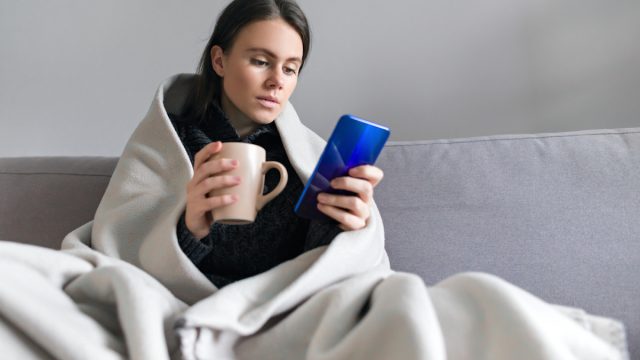 young woman wrapped in blanket with mug, looking at phone