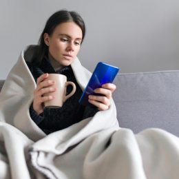 young woman wrapped in blanket with mug, looking at phone