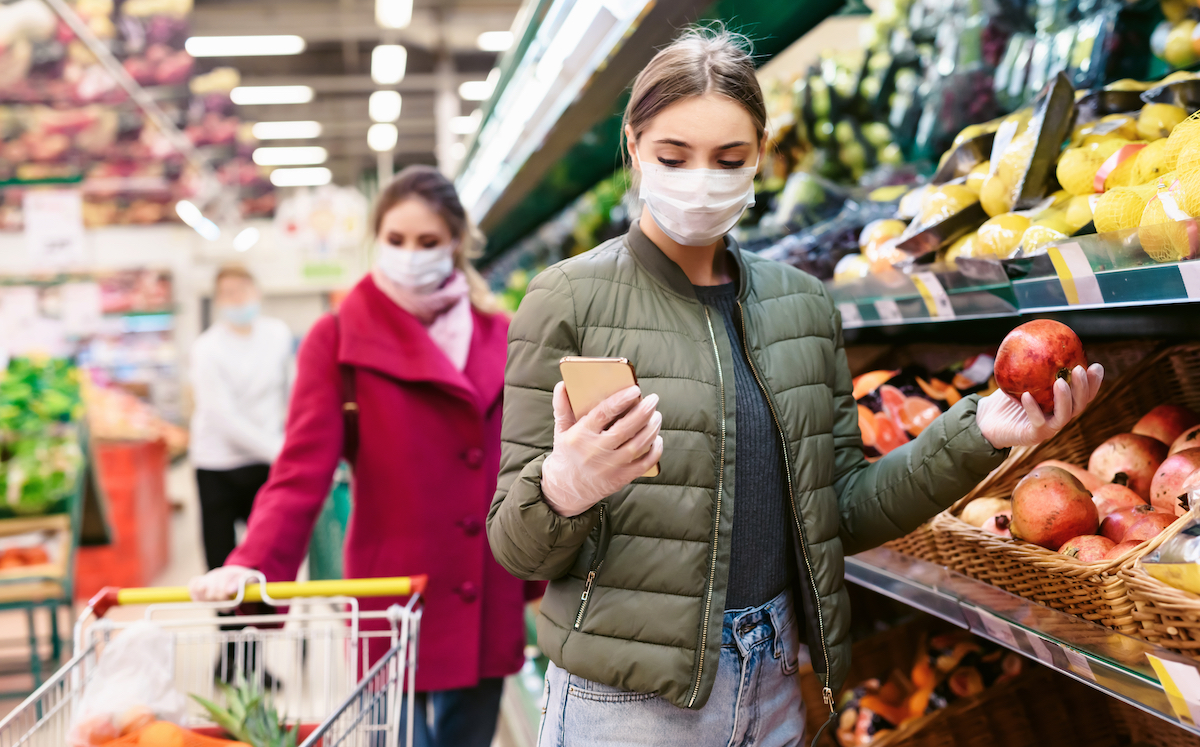 A young woman in a disposable face mask is checking a shopping list on a smartphone while there is another woman with shopping cart background