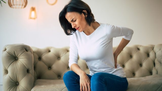 Axial pain. Close-up photo of a hurting woman, who is sitting on a couch and holding her lower back with her left hand.