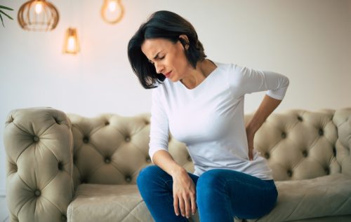 Close-up photo of a hurting woman, who is sitting on a couch and holding her lower back with her left hand.