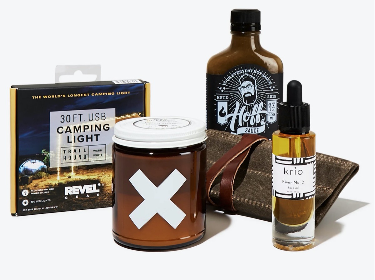 camping lights, brown glass encased candle with x on it, face oil, leather charging cable roll, and bottle of hot sauce on white background