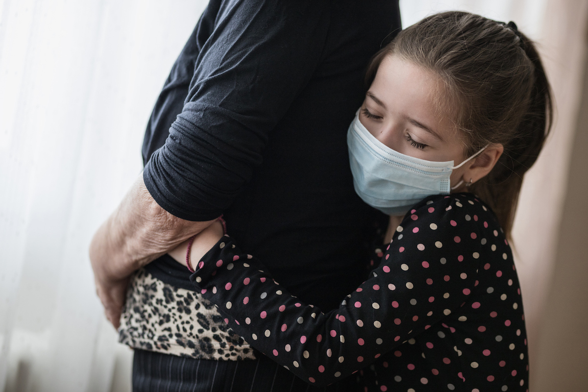 Young child with her eyes closed and protective mask on giving her grandparent a hug from behind