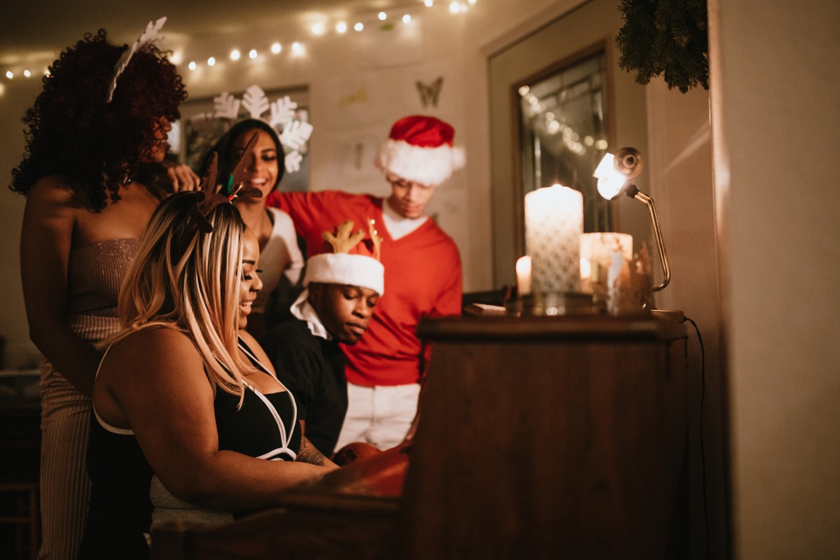 A group of young adult friends gather at a home for Christmas celebration over the holiday, dressed to fit the occasion with various Christmas accessories. They sing songs together at the piano, enjoying the Christmas cheer.