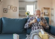 Senior man suffering from flu drinking tea while sitting wrapped in a blanket on the sofa at home. Sick older man with headache sitting under the blanket in the living room. Man with a cold lying on the sofa holding a mug of hot tea.