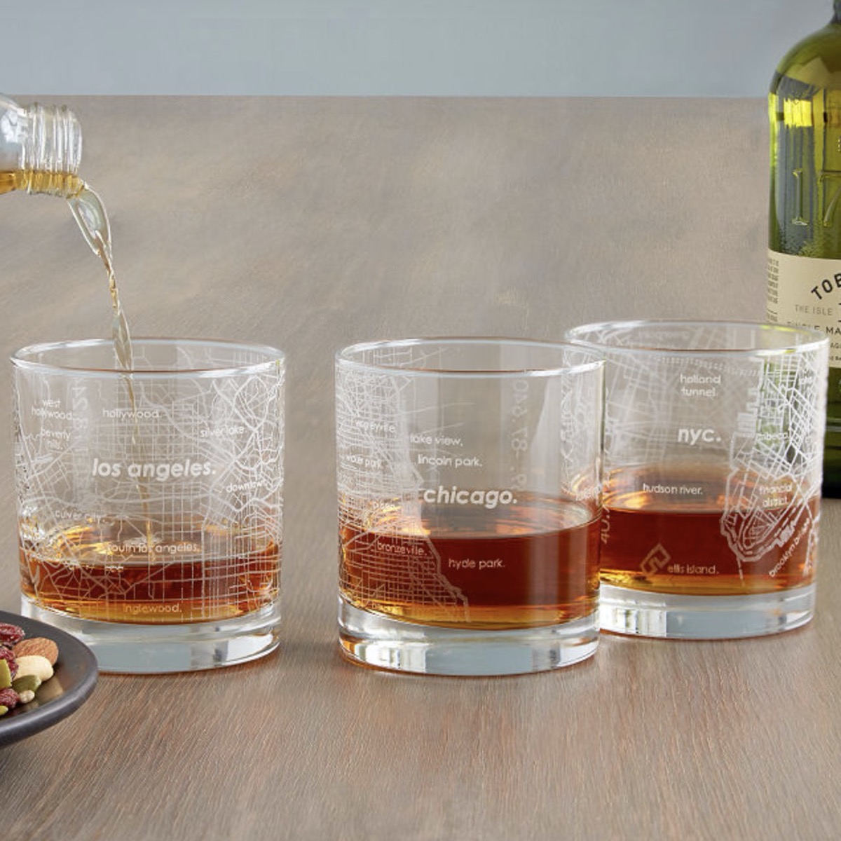 bottle pouring dark liquor into a set of three etched glasses with maps of los angeles, chicago, and nyc etched into them