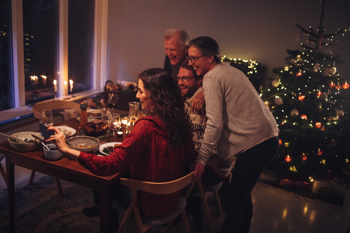 Four people at a table in front of a Christmas tree and one woman holding a phone