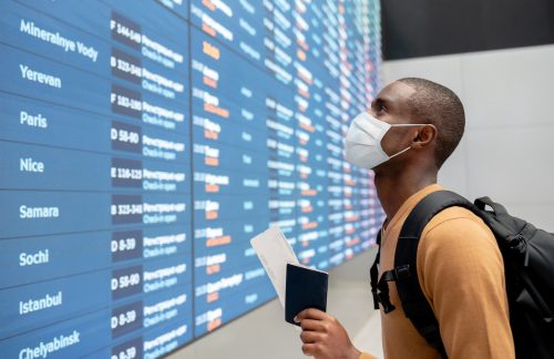 ภาพเหมือนของนักเดินทางชายสวมหน้ากากอนามัยที่สนามบินและมองดูตารางเที่ยวบิน
