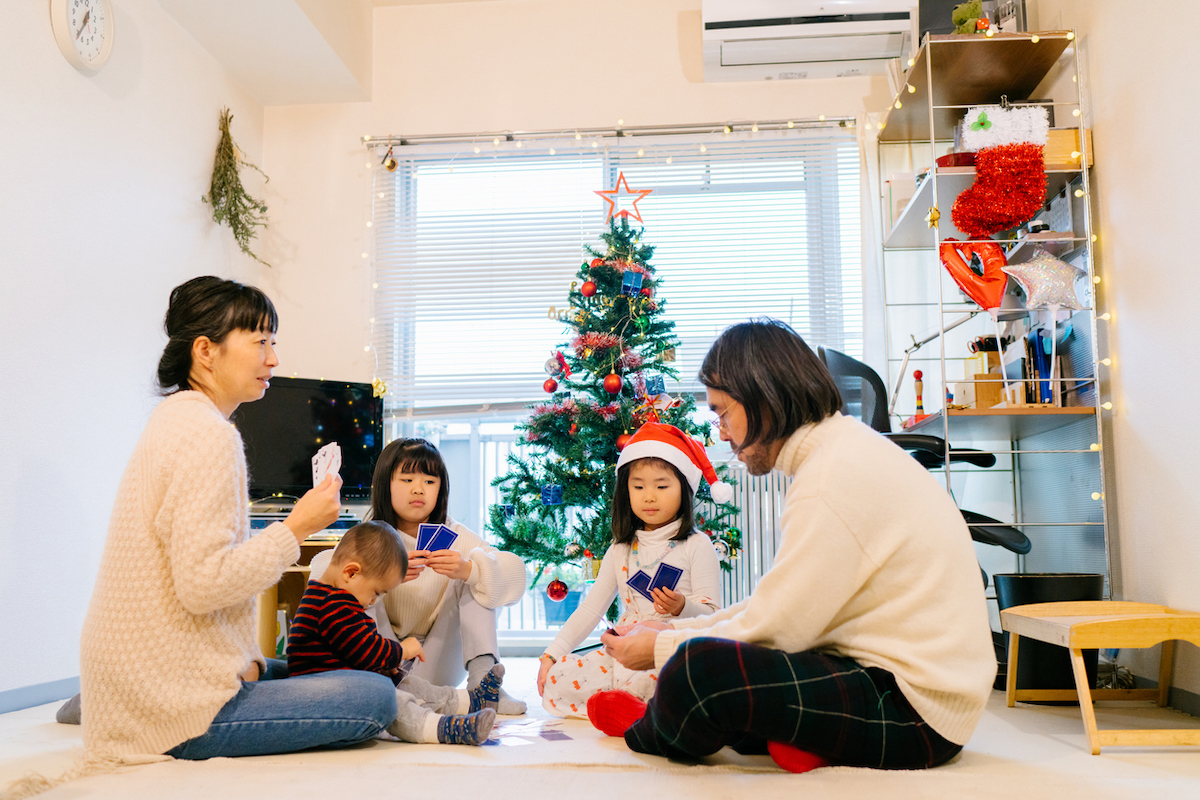 A family is sitting on floor and playing cards games in the living room decorated with Christmas ornaments and a Christmas tree during Christmas.
