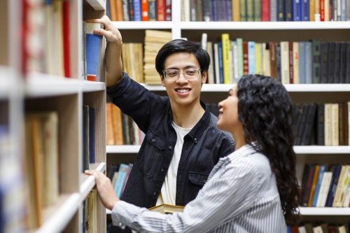 young man in glasses taking book from library bookshelf in campus library while smiling at girl
