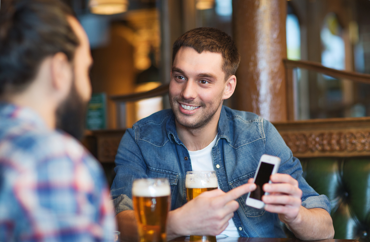 male holds up his smartphone while drinking a beer at a bar