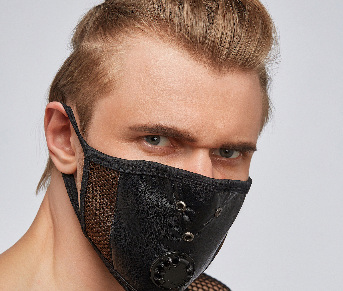 Close-up portrait of a blond man, wearing a black leather mask with brown mesh under it and ventilation openings.