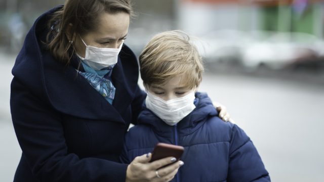 A mother wearing a face mask shows her son something on her smartphone while he also wears a mask.