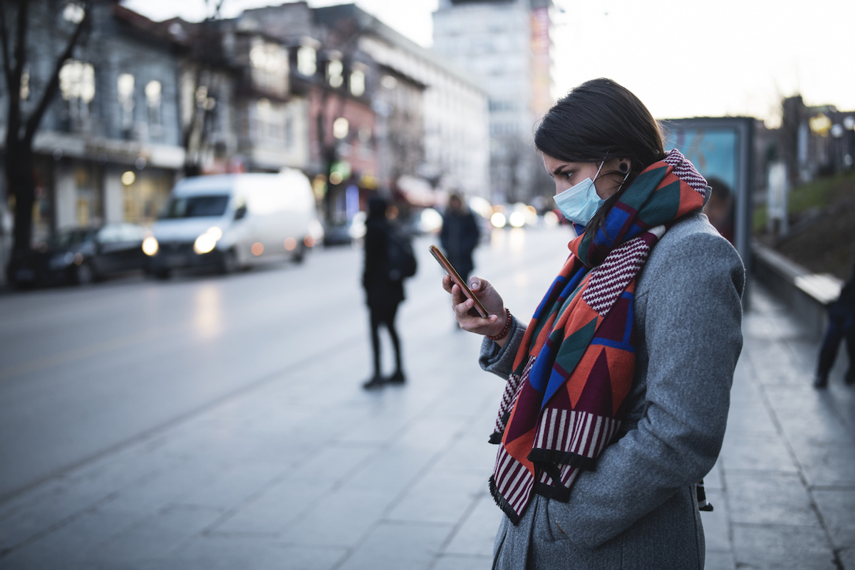 Side view of young woman with face mask on the street, waiting for city transport and looking down at her smartphone.