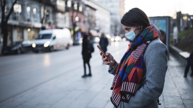 Side view of young woman with face mask on the street, waiting for city transport and looking down at her smartphone.