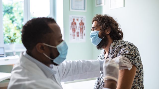 doctor in surgical mask giving male patient in mask a vaccine in arm