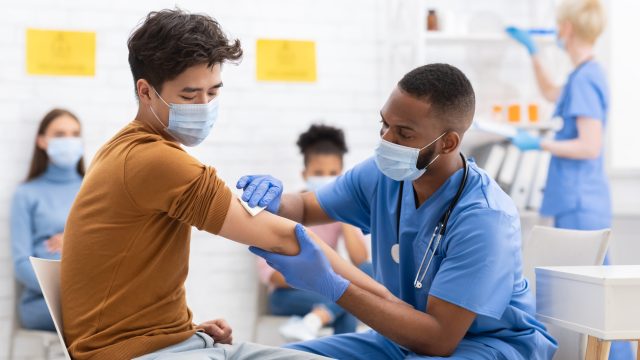 A young male patient wearing a face mask prepares to receive a coronavirus vaccine from a young male healthcare worker.