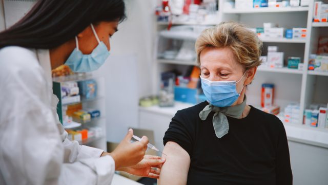 A senior woman wearing a face mask receives a COVID vaccine from a young female pharmacist who is also wearing a face mask.