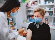 A senior woman wearing a face mask receives a COVID vaccine from a young female pharmacist who is also wearing a face mask.