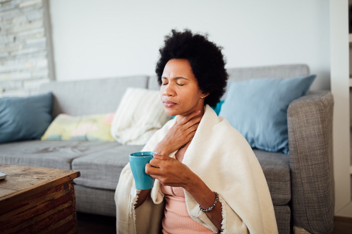 woman, fallen ill is staying at home wrapped in a blanket socially distancing and quarantining herself, feeling her throat hurt and being sore, having a cup of hot tea