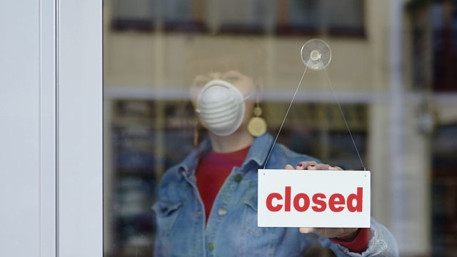 A woman wearing a face mask turns around a closed sign on the door of a business that was forced to shutter due to COVID