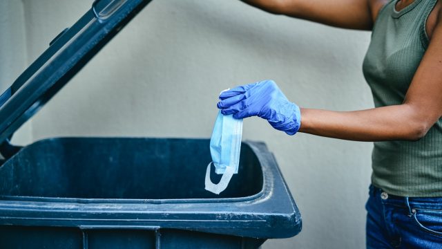 A young woman wearing protective gloves throwing away a face mask into a garbage can.