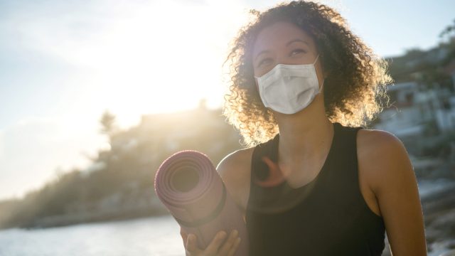 A young woman wearing a face mask outdoors while holding a yoga mat.