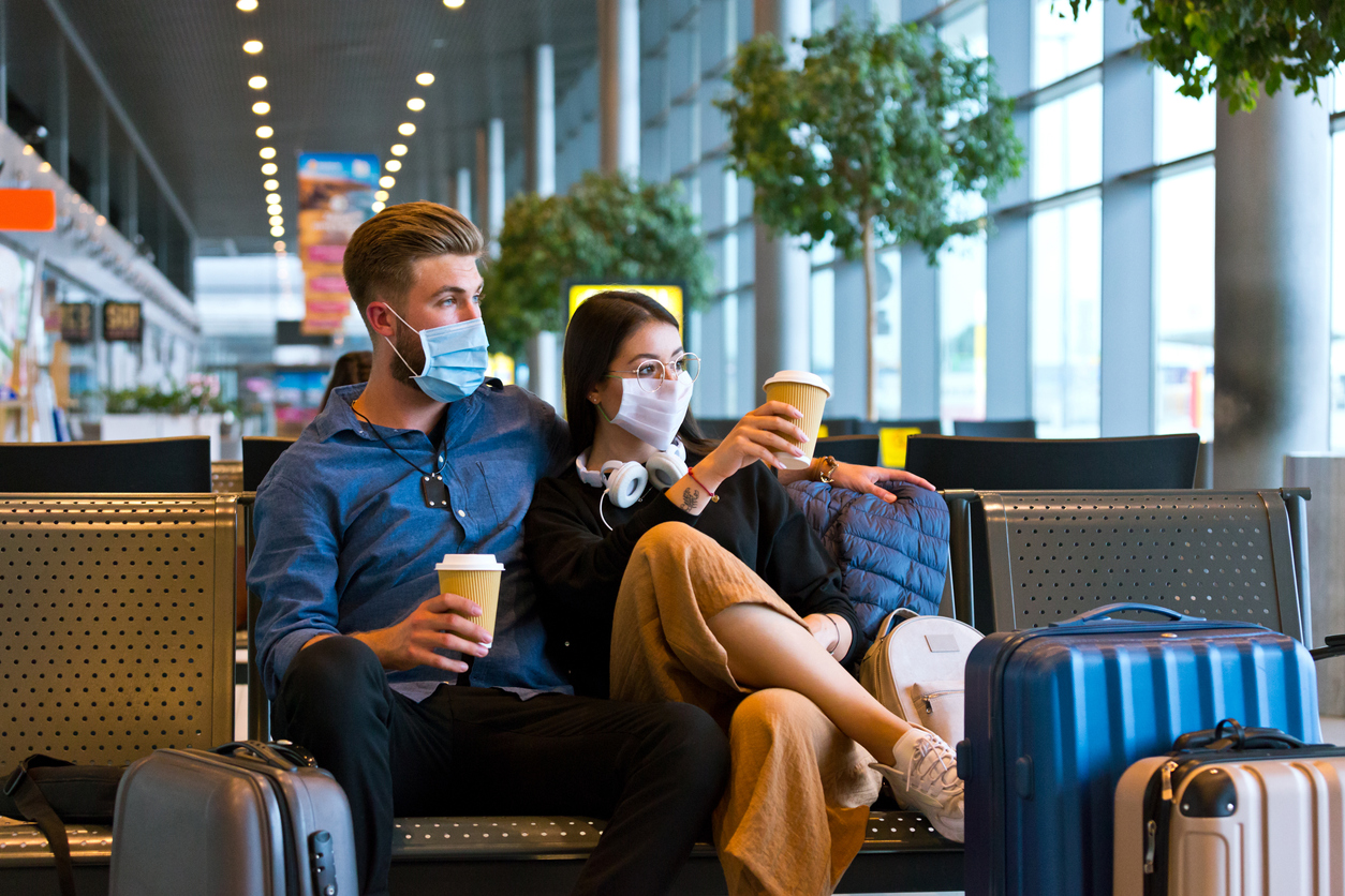 A young couple wearing face masks while sitting in a transit lounge waiting to travel during the holidays.
