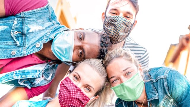 Millenial friends taking selfie smiling behind face masks – Happy friendship and new normal concept with young people having fun together – Bright sunshine filter with focus on left girl