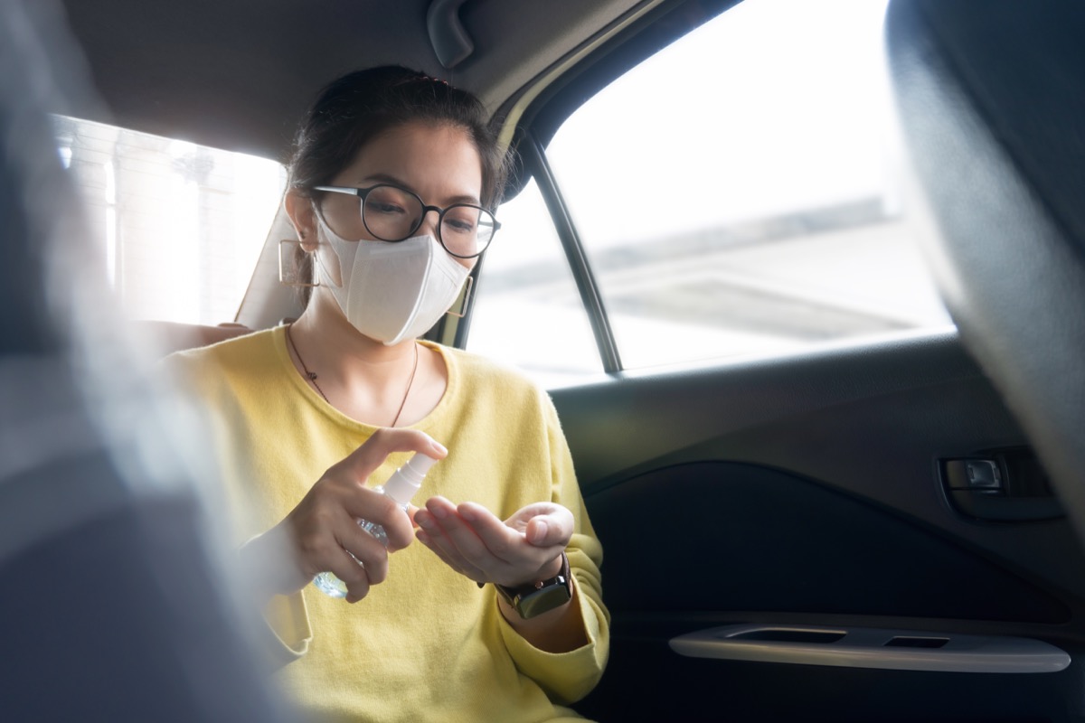 Woman with protective mask spraying disinfectant alcohol on her palms and her hands to prevent coronavirus in a car