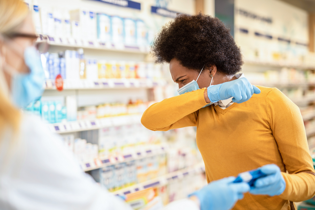 woman coughing while wearing a mask and asking for medication at a pharmacy