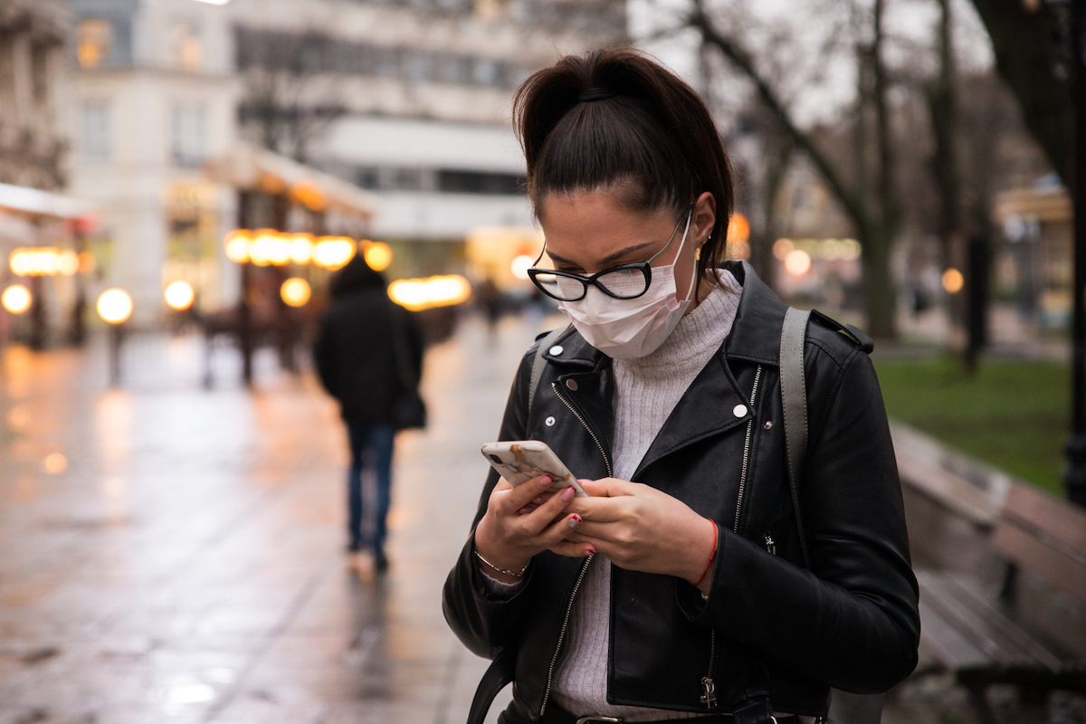 Side view of young woman with face protective mask in a city at dusk, she is using smartphone.