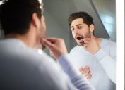 Handsome young man with stubble keeping mouth open while checking tooth and looking into mirror in bathroom