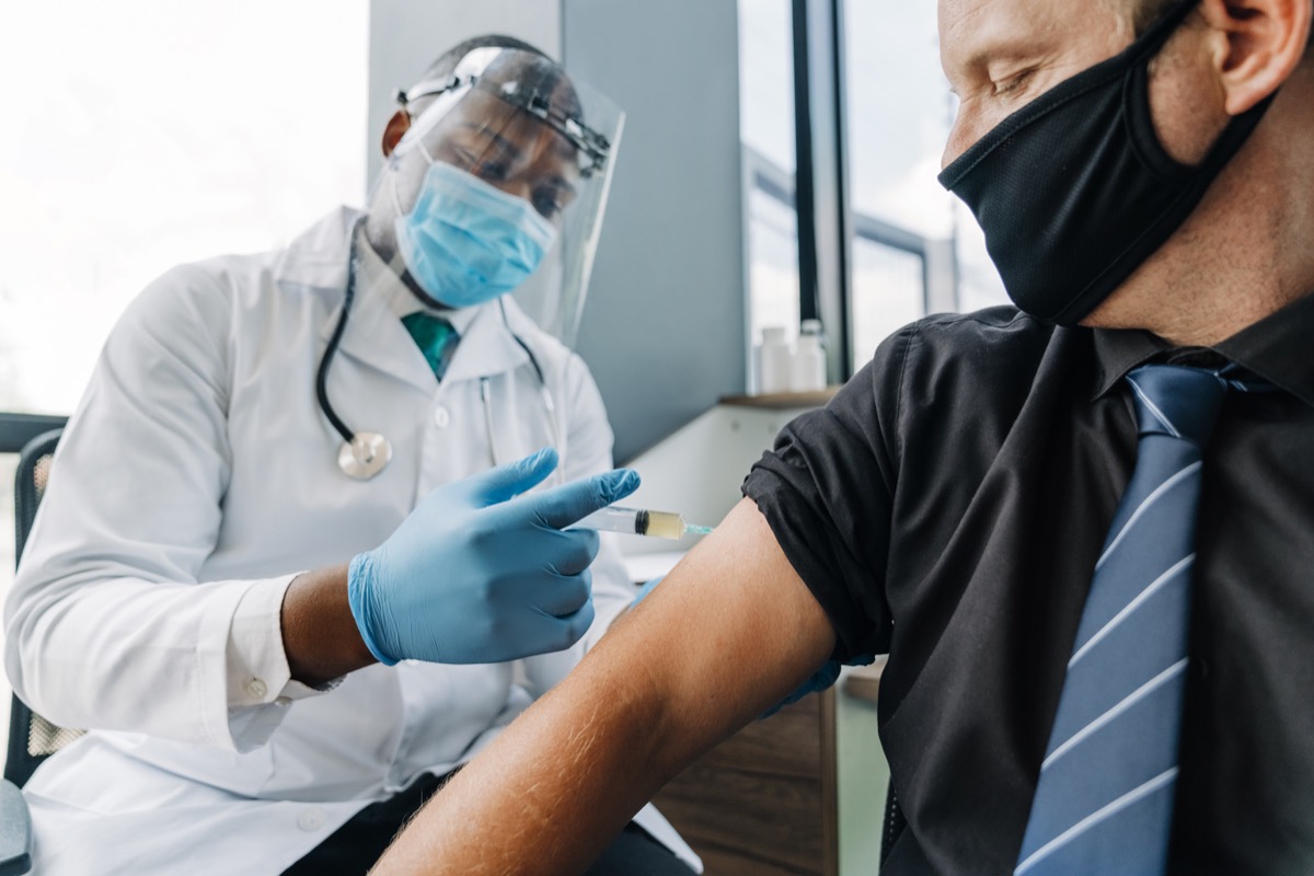 Doctor injecting vaccine on male patient's arm. Man is wearing face mask while sitting with healthcare worker. They are at medical clinic during coronavirus.