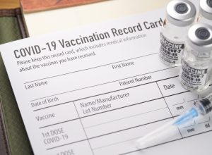 Vials of COVID-19 vaccine and a syringe sit on top of a vaccination date record card.