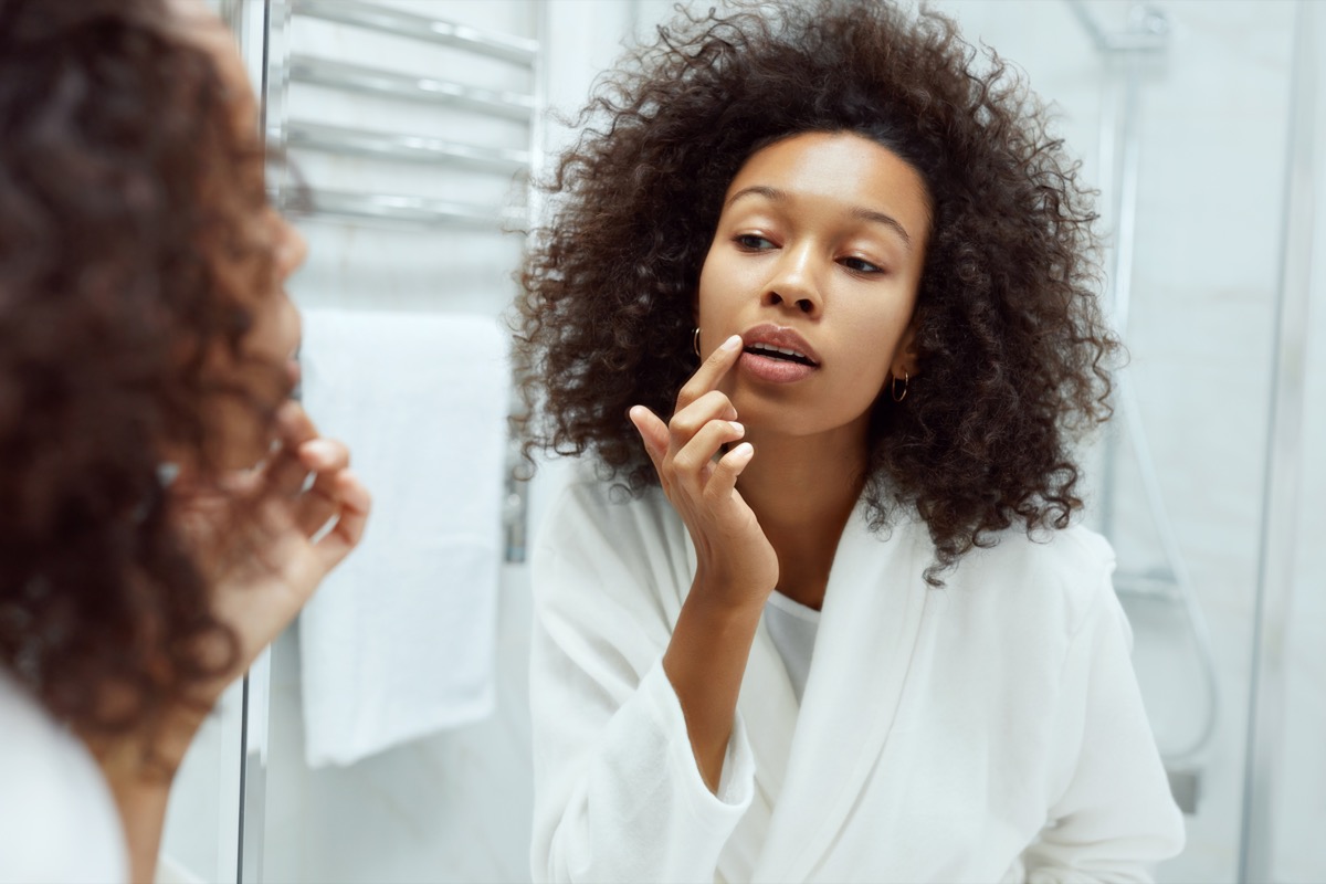 Lips skin care. Woman applying lip balm looking in mirror at bathroom. Portrait of beautiful african girl model with beauty face and natural makeup applying lip product with finger