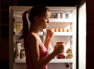 Young woman eating ice cream near refrigerator at night